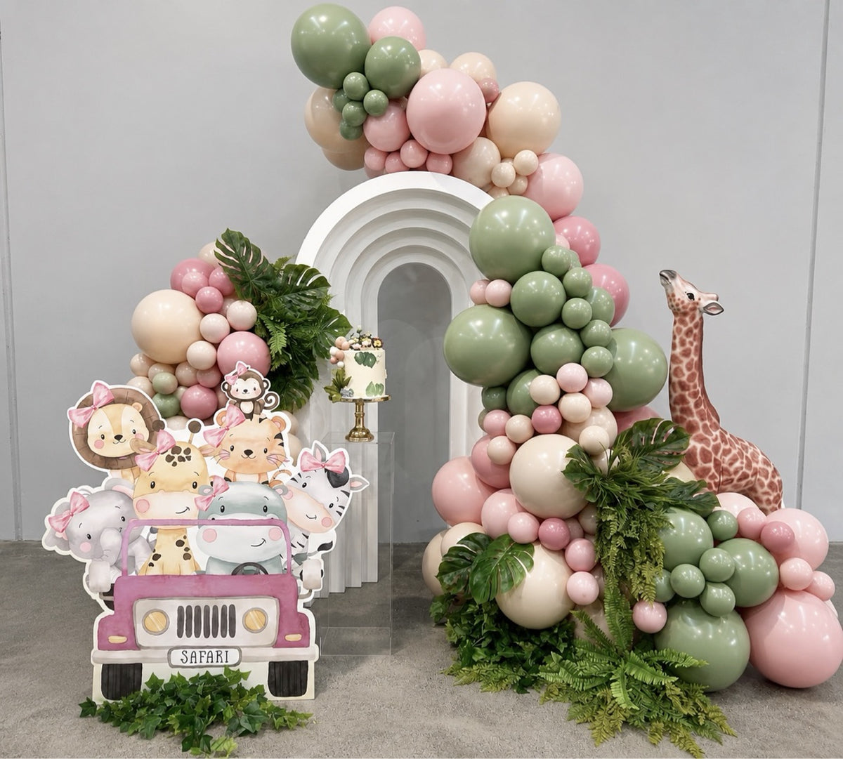 Pink safari backdrop with white open arch, blush pink and sage balloon garlands, tropical leaves and a safari jeep cutout with baby animals