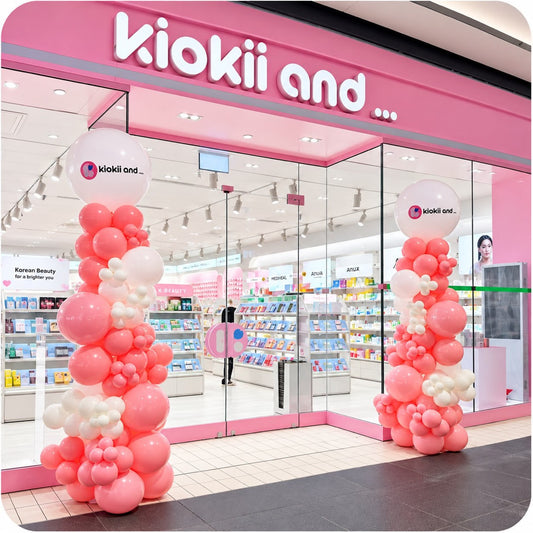 Two organic balloon columns / pillars in pink and white with Kiokii And logo toppers at a K-beauty store entrance in a mall