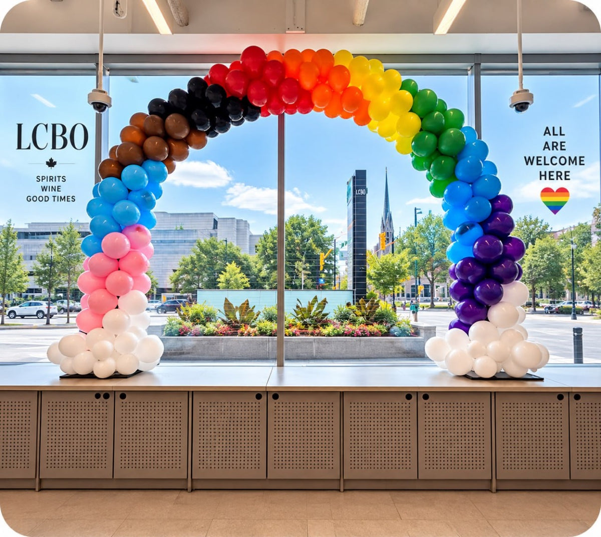 Rainbow balloon arch in Pride colors inside an LCBO store with an ALL ARE WELCOME HERE window sign