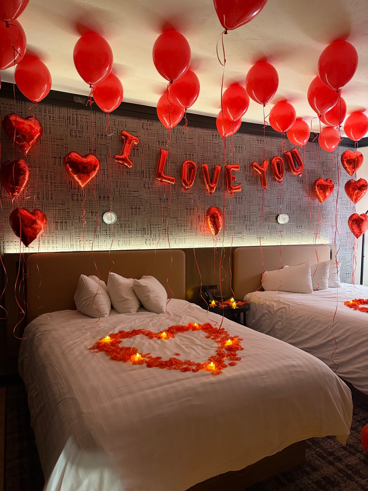 Hotel bedroom with I LOVE YOU red foil letter balloons, red heart balloons and a heart of rose petals with candles on the bed