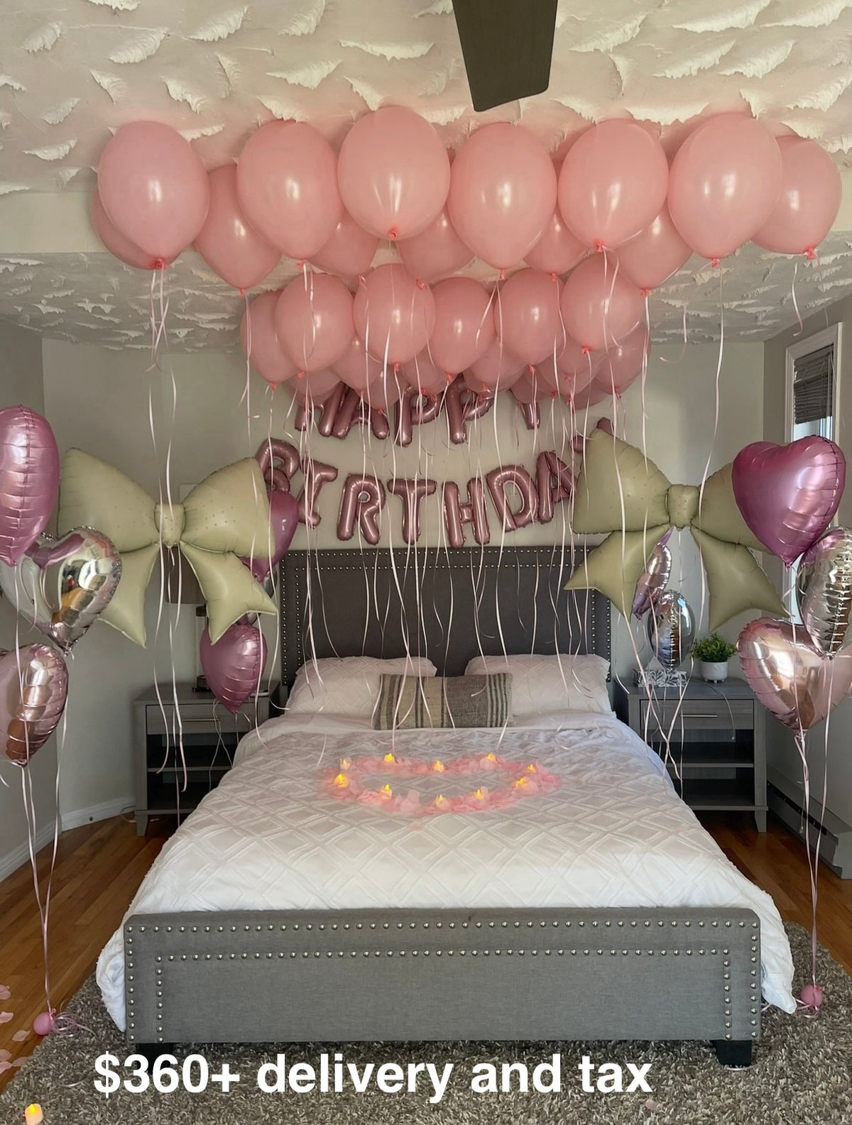 Pink birthday bedroom setup with helium balloons on the ceiling, Happy Birthday foil letters, giant bow balloons and a heart of rose petals with candles on the bed