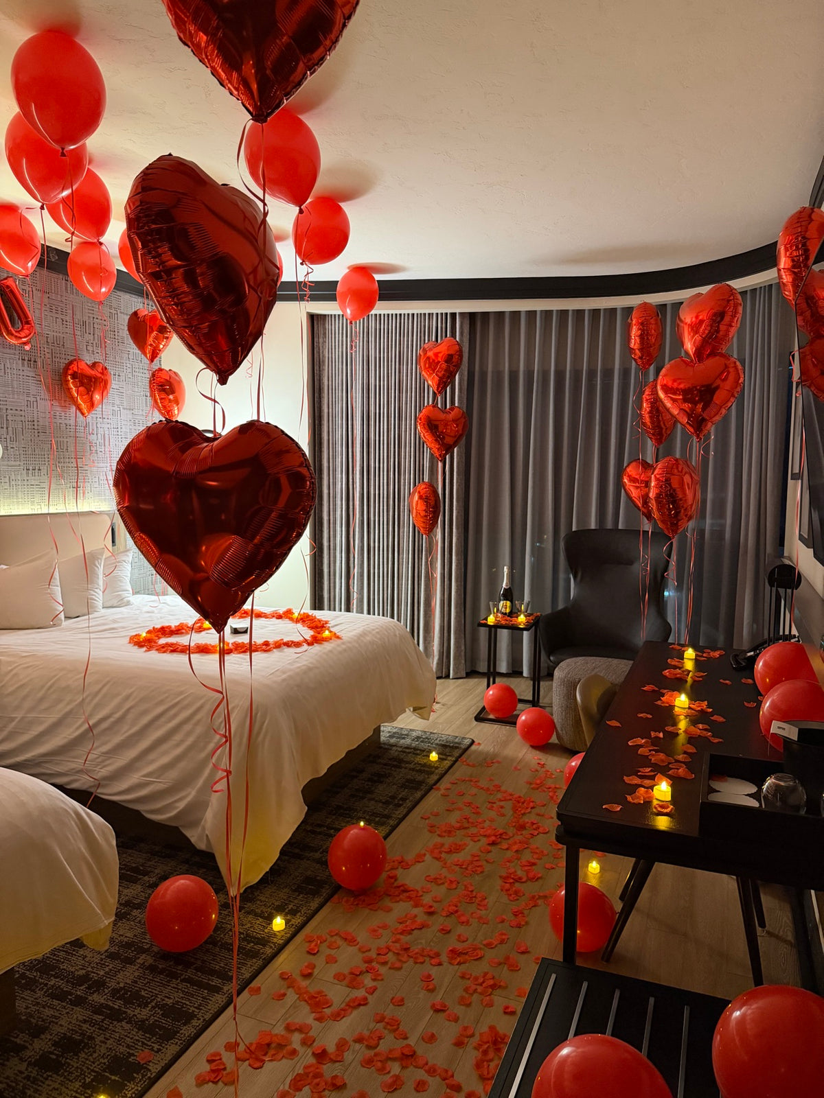 Romantic hotel room decorated with red helium balloons on the ceiling, giant red heart foil balloons, rose petals and candles on the bed