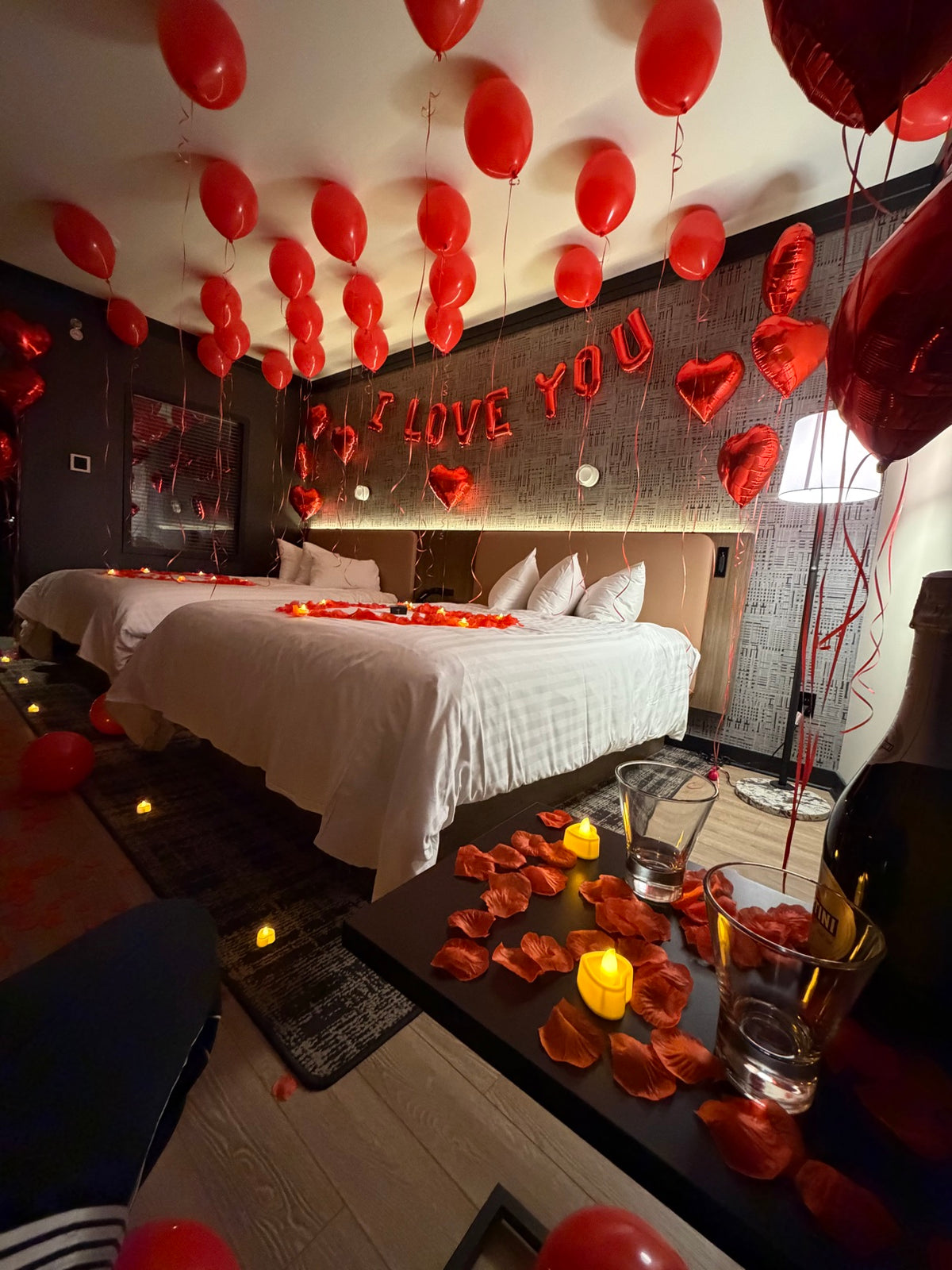 Hotel room entrance decorated with red heart foil balloon columns, rose petals and candles leading to the bed