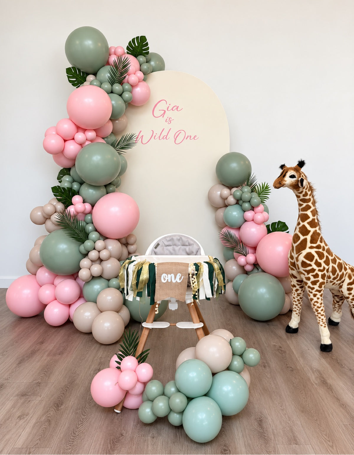 Wild One safari first birthday backdrop with cream arch panel, personalized name text, blush pink and sage green balloon garlands and tropical leaves